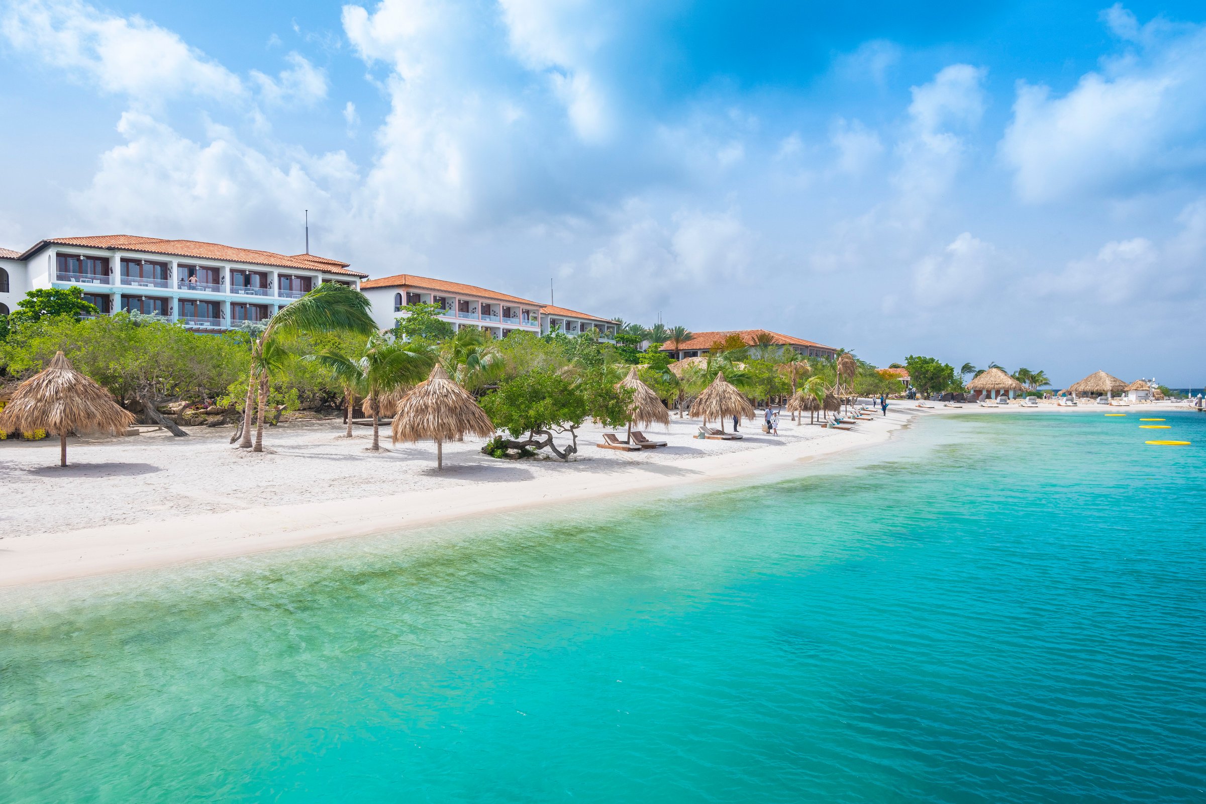 White-sand beach with palapas, resort buildings and calm turquoise water.