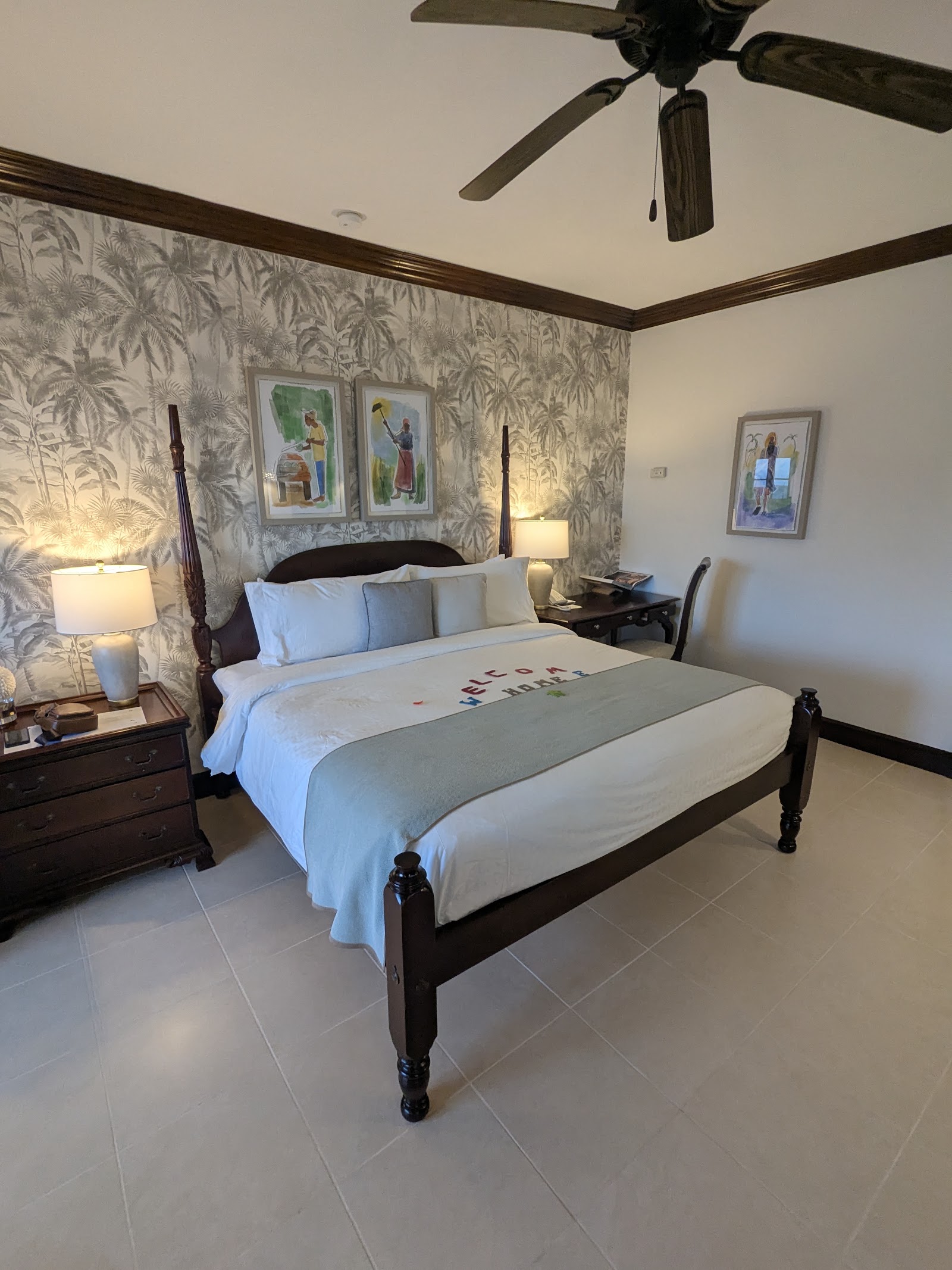 Resort bedroom with four-poster bed, palm-patterned wallpaper, bedside lamps, dark wood furniture, and ceiling fan.