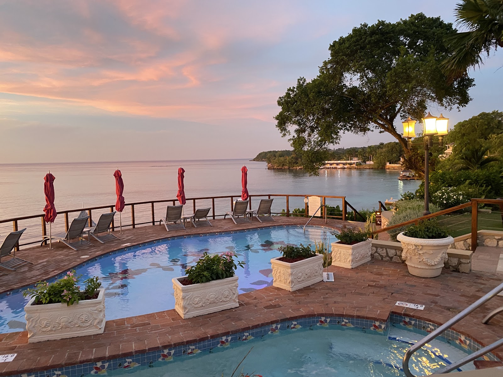 Waterfront pool deck at Sandals Royal Plantation with lounge chairs, red umbrellas, and calm bay waters at dusk.