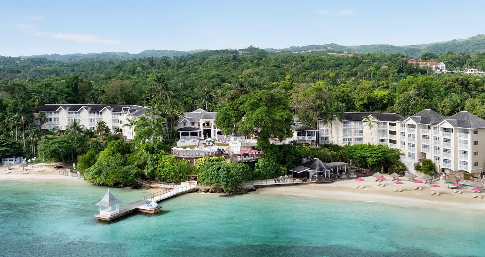 White resort buildings nestled among tropical vegetation overlook a turquoise beach with pink umbrellas and a wooden pier extending into clear water.