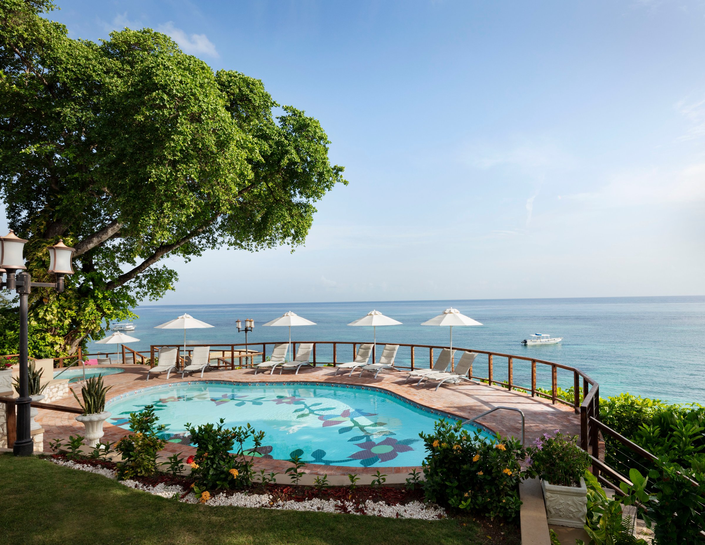 Mosaic pool on a garden lawn with umbrellas and a sea-view railing.