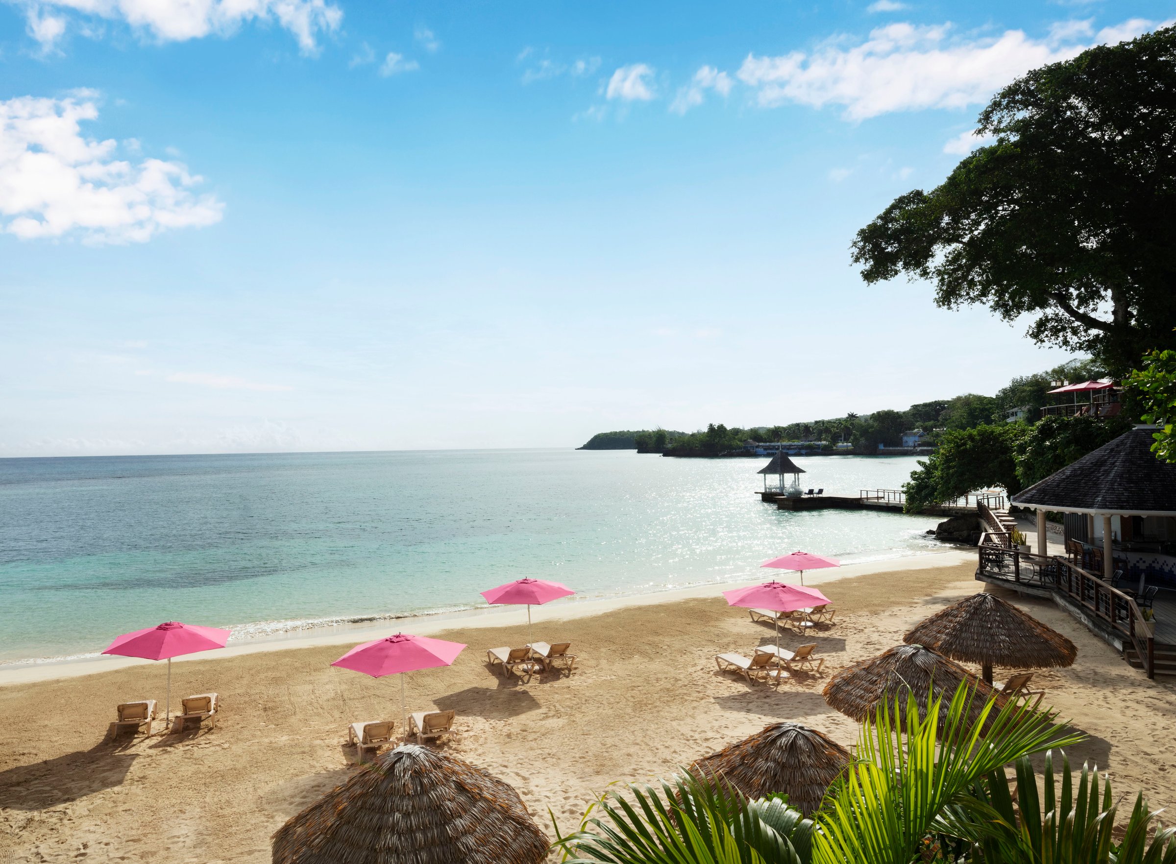 Private beach with pink umbrellas, thatched shades and loungers on a calm bay.