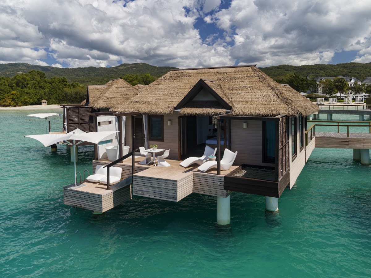 Overwater bungalow with thatched roof and wooden deck surrounded by turquoise water, forested hills beyond.
