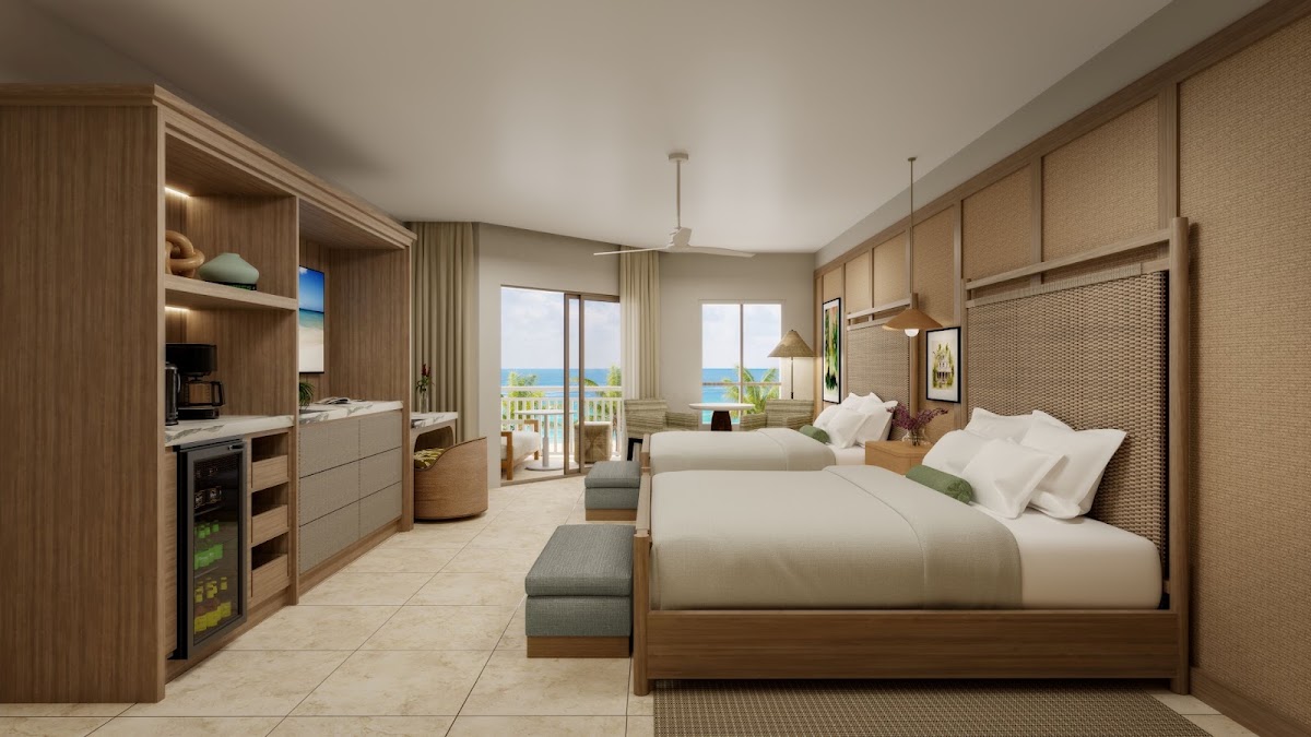 A modern resort room with two queen beds, wood furnishings, kitchenette, and ocean views through floor-to-ceiling balcony doors at Sandals South Coast.