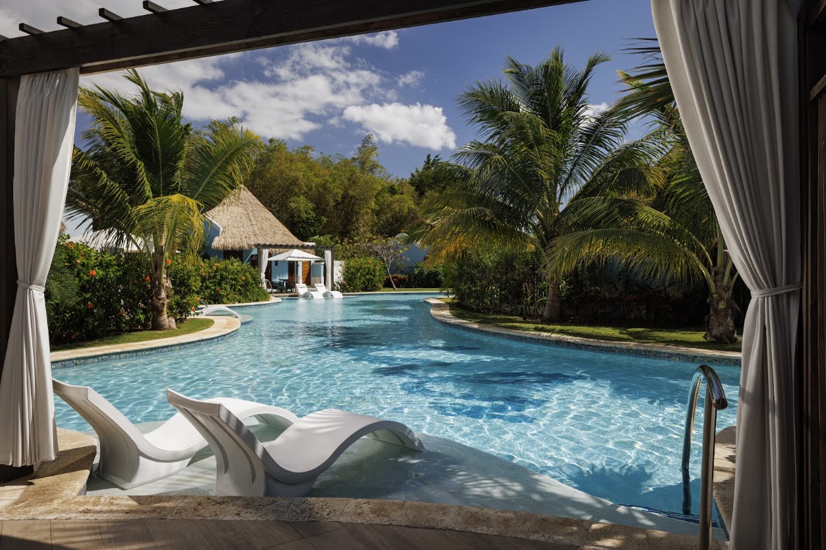 White lounge chairs on the pool deck at Sandals South Coast, with a lagoon-style pool, palm trees, and a thatched-roof bungalow visible beyond.