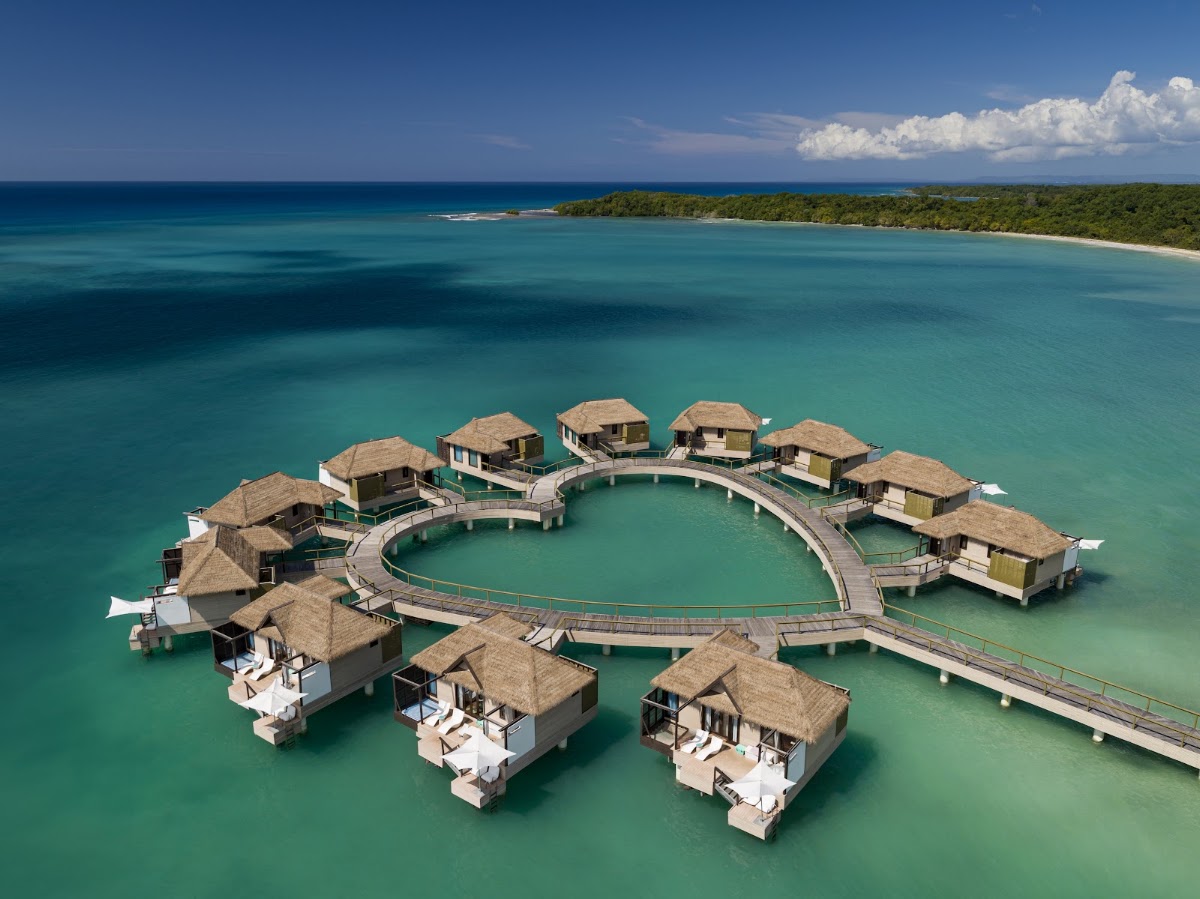 Aerial view of thatched-roof overwater bungalows arranged in a circle connected by wooden walkways in turquoise lagoon water, with forested coastline beyond.