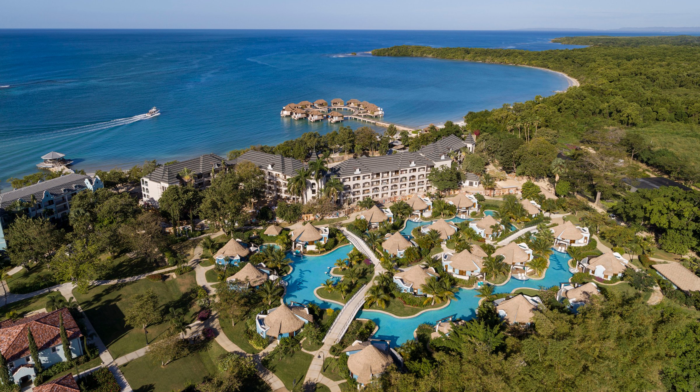 Aerial of thatched rondovals around the river pool toward the over-water villas.