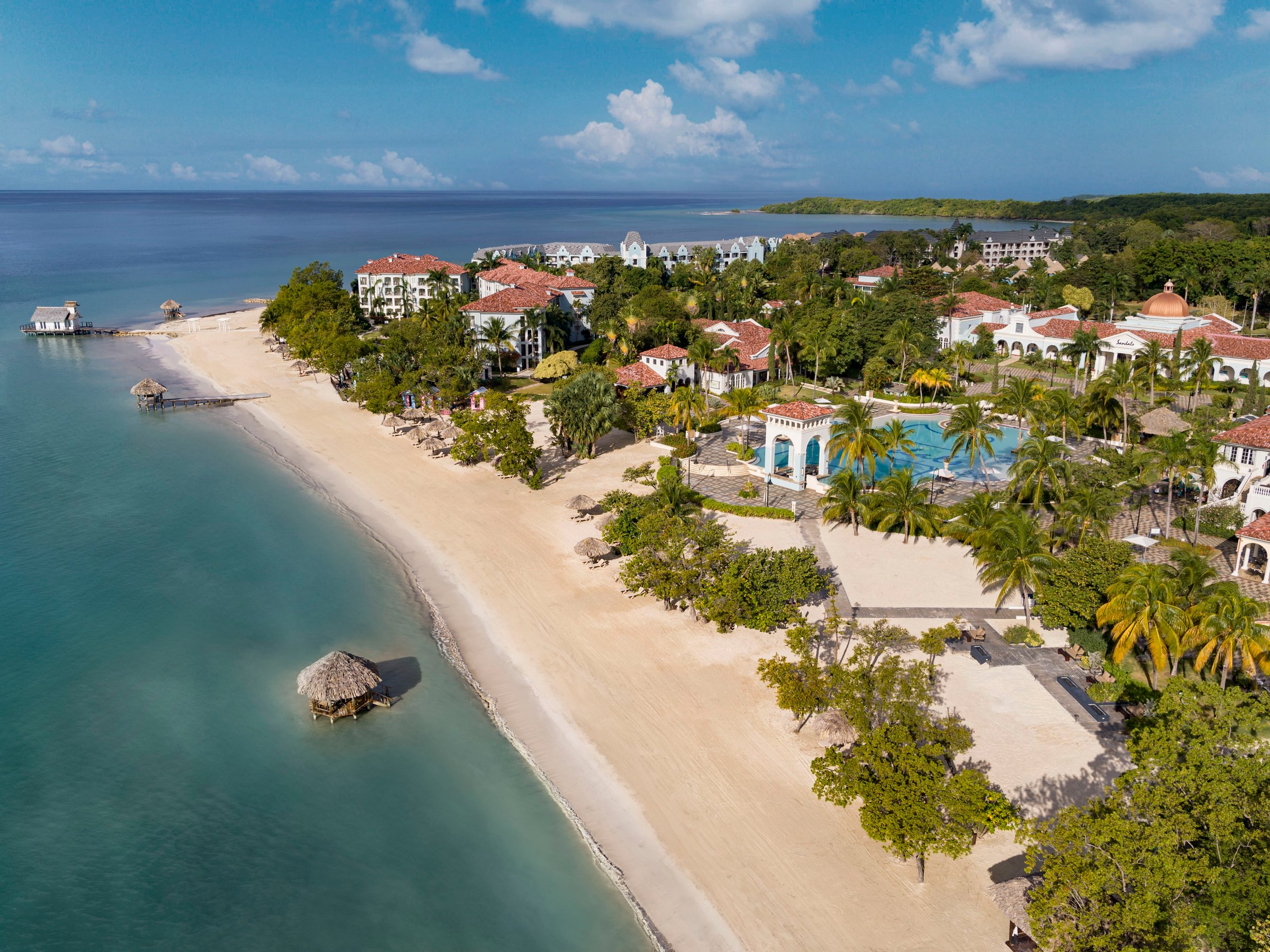 Aerial along the long sandy beach, piers and red-roofed resort.
