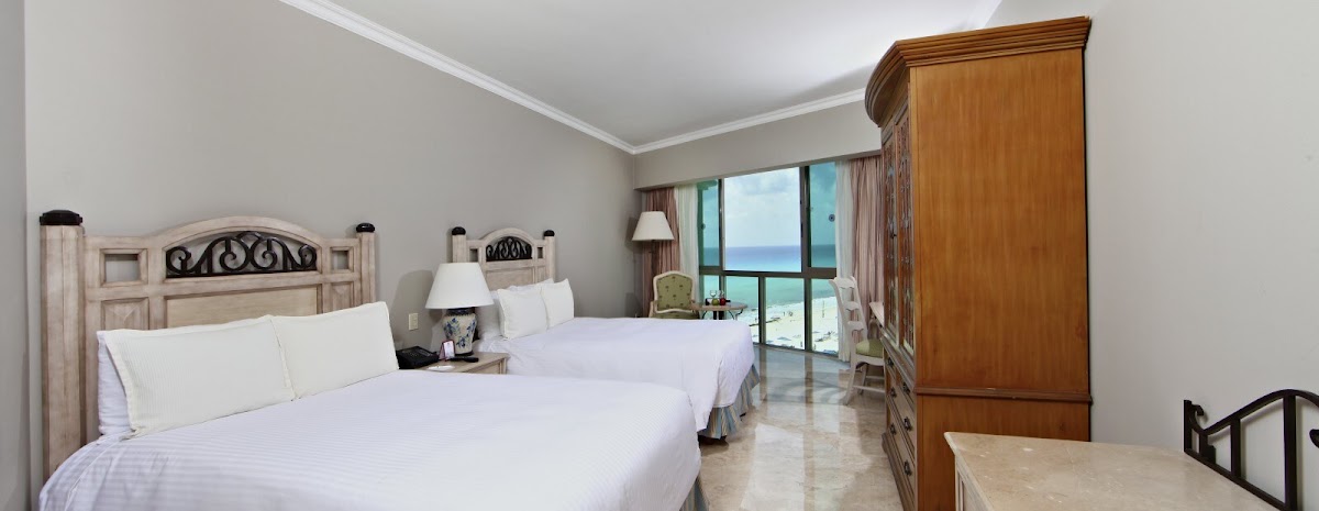 Resort guest room with two double beds, wooden furniture, and ocean view through balcony doors at Sandos Cancun.