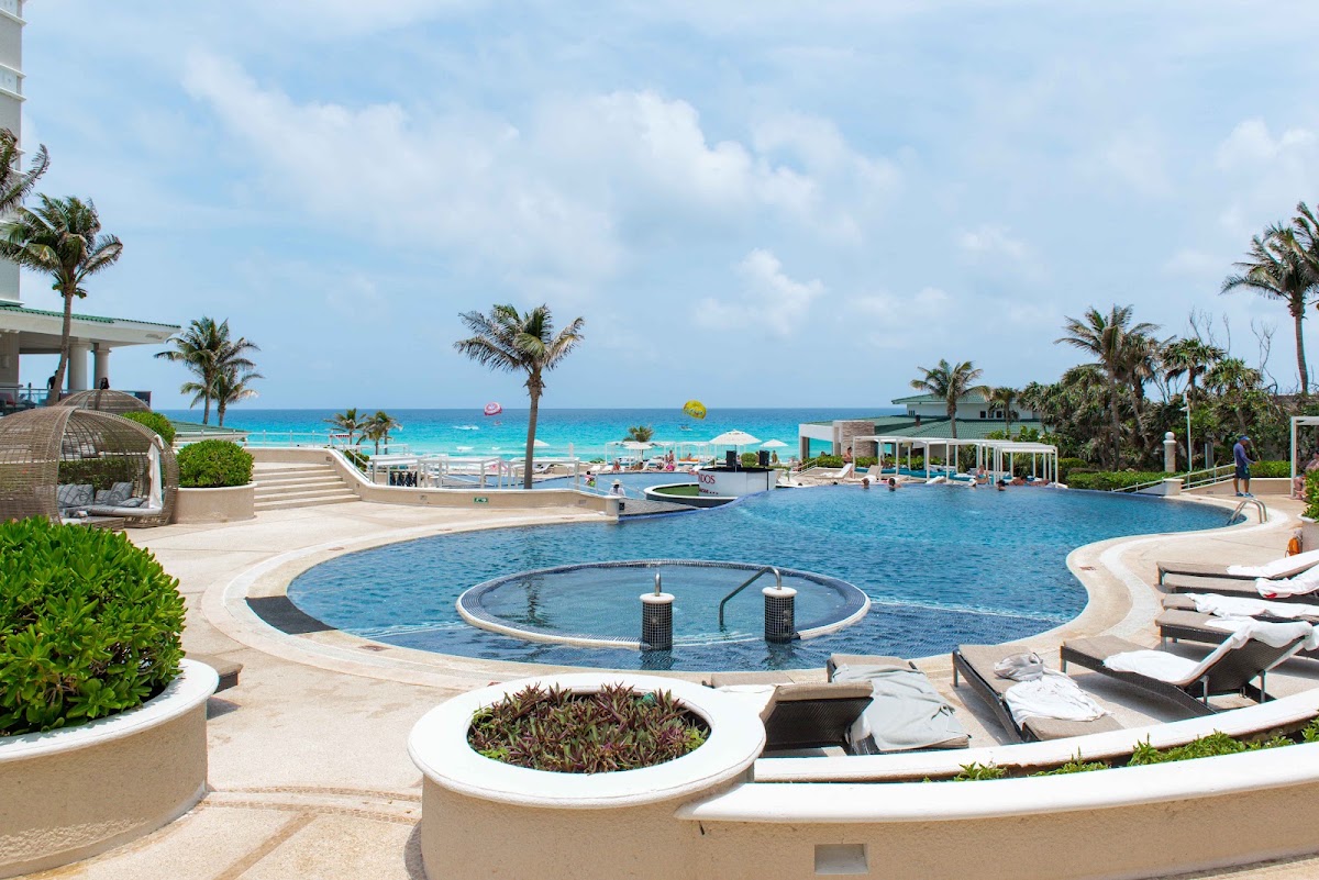 Resort pool with circular hot tub in foreground, beach and turquoise Caribbean Sea beyond, palm trees and lounge chairs throughout.