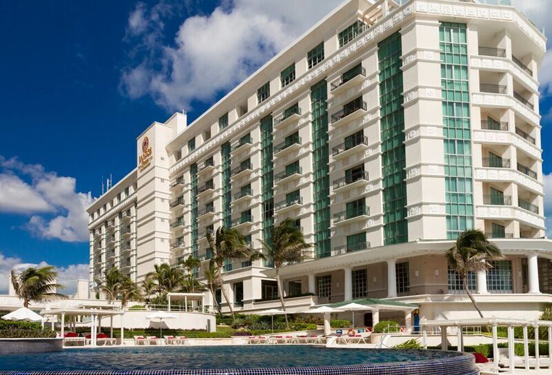 White multi-story beachfront hotel with green-trimmed balconies, palm trees, and waterfront lounge chairs under blue sky.