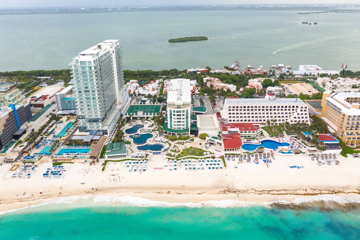 Aerial view of Sandos Cancun resort with beachfront towers, multiple pools, sandy beach, turquoise water, and adjacent hotels along the coast.