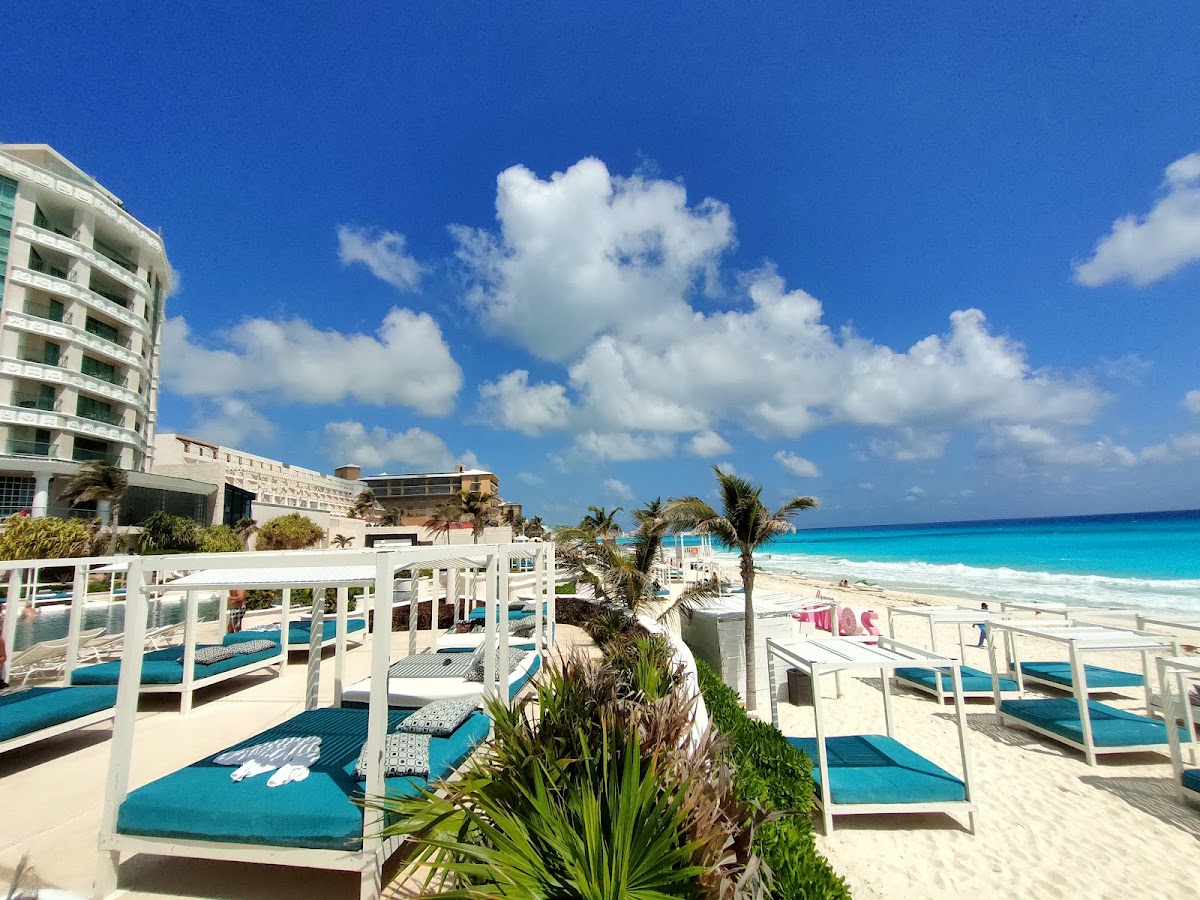 White cabana beds with turquoise cushions line the sandy beach at Sandos Cancun, with palm trees and Caribbean Sea beyond.