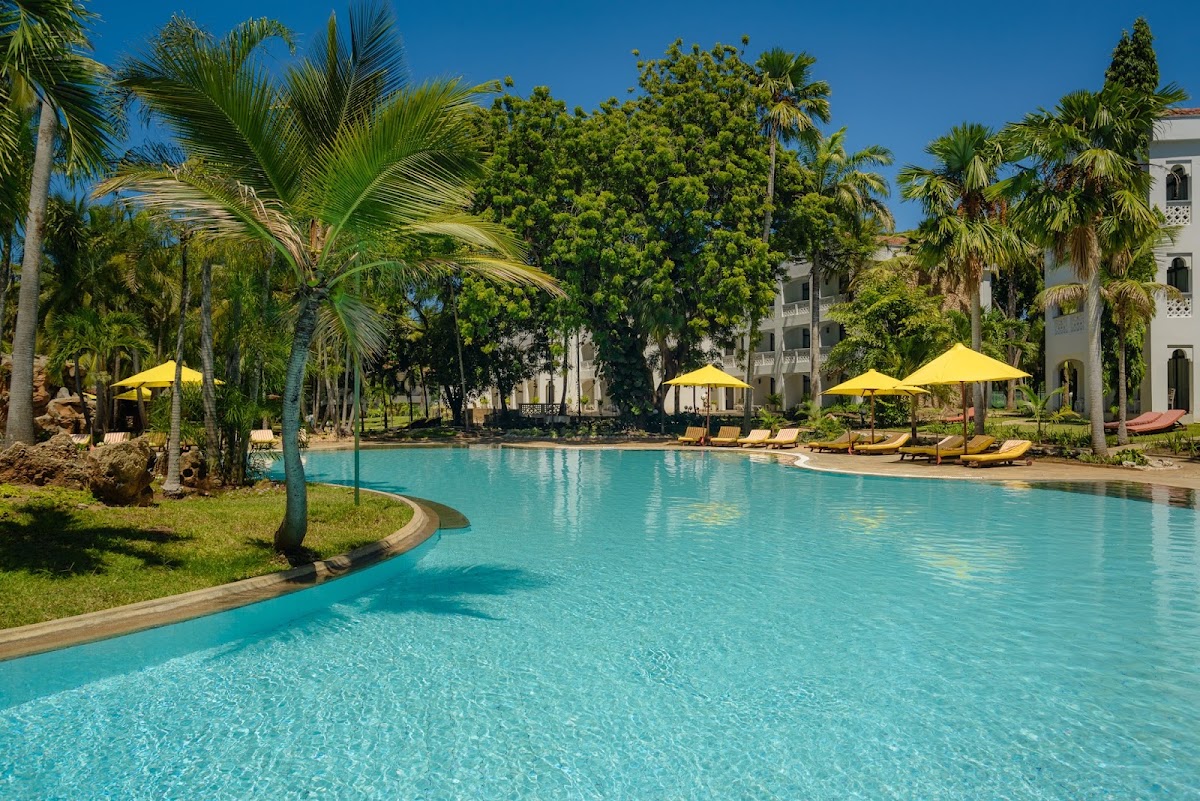 Turquoise pool surrounded by palm trees and yellow umbrellas with lounge chairs at Sarova Whitesands Beach Resort, white building visible behind.