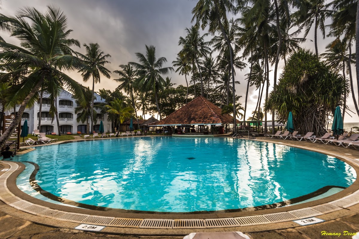 Large curved swimming pool surrounded by palm trees, lounge chairs, and a thatched-roof pavilion at Sarova Whitesands Beach Resort.