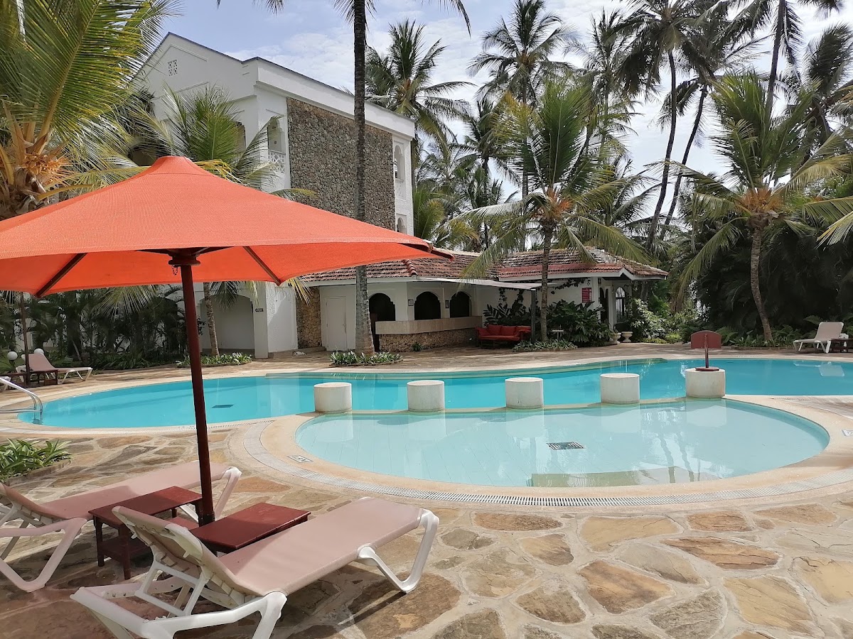 Sarova Whitesands Beach Resort pool area with red umbrella, lounge chairs, circular hot tub, and palm trees surrounding white colonial-style buildings.