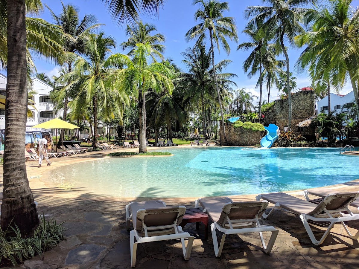 Lounge chairs line the sandy beach at Sarova Whitesands, with turquoise lagoon water, palm trees, and a blue water slide visible beyond.
