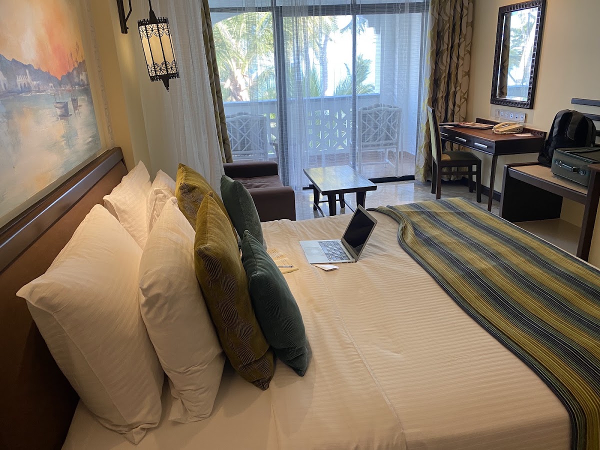 Spacious resort bedroom with king bed, balcony overlooking palm trees and ocean, work desk, and sitting area with ocean view.