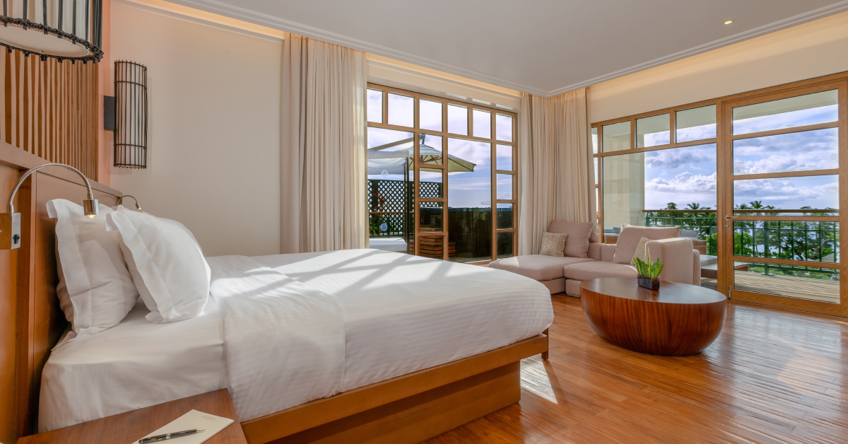 Spacious resort bedroom with wood bed, white linens, lounge seating, and ocean views through glass doors opening to a coastal balcony.