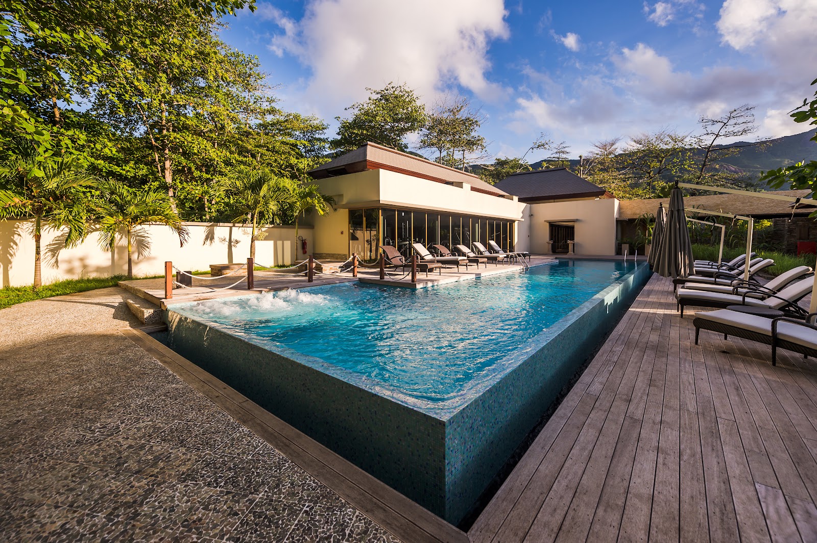 Resort pool with turquoise water surrounded by lounge chairs, wooden deck, modern white buildings, and lush tropical vegetation against mountains.