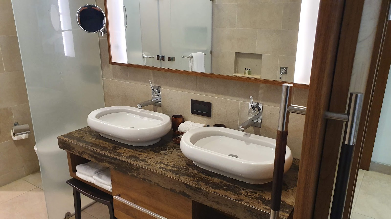 Bathroom vanity with two white oval sinks on a dark wood countertop, illuminated mirror, and beige tile walls at Savoy Seychelles Resort & Spa.