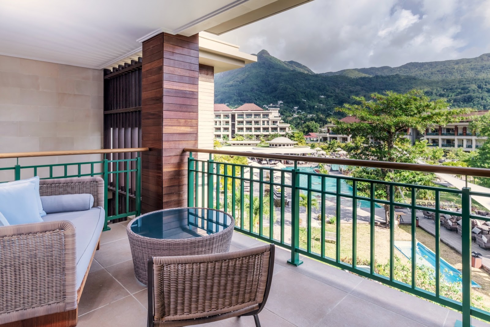 Balcony seating area with wicker furniture and mountain views over a resort with lush green hills beyond.