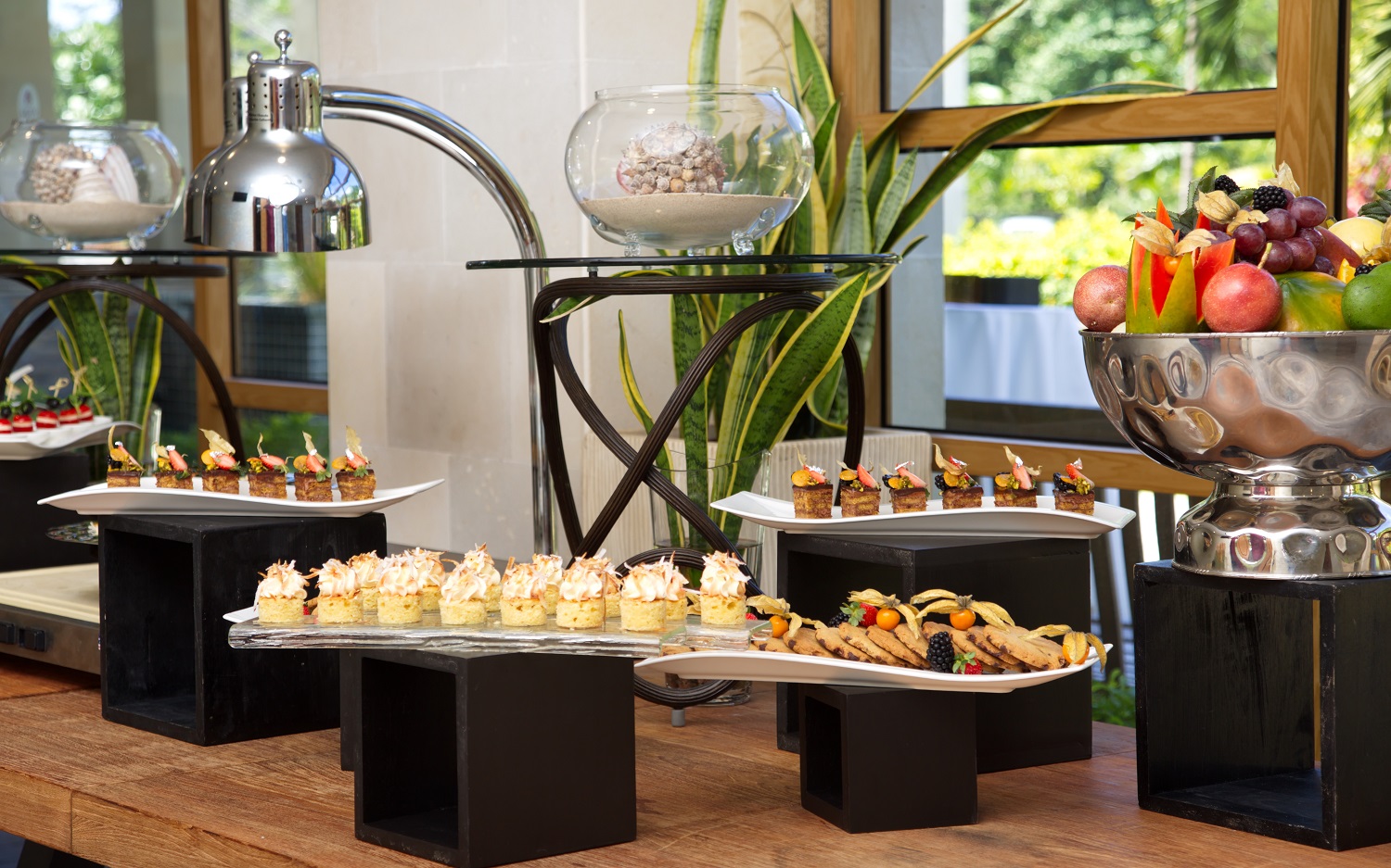 Indoor buffet display at Savoy Seychelles with appetizers, pastries, fresh fruit, and metal serving pieces on black pedestals beside potted plants and large windows.