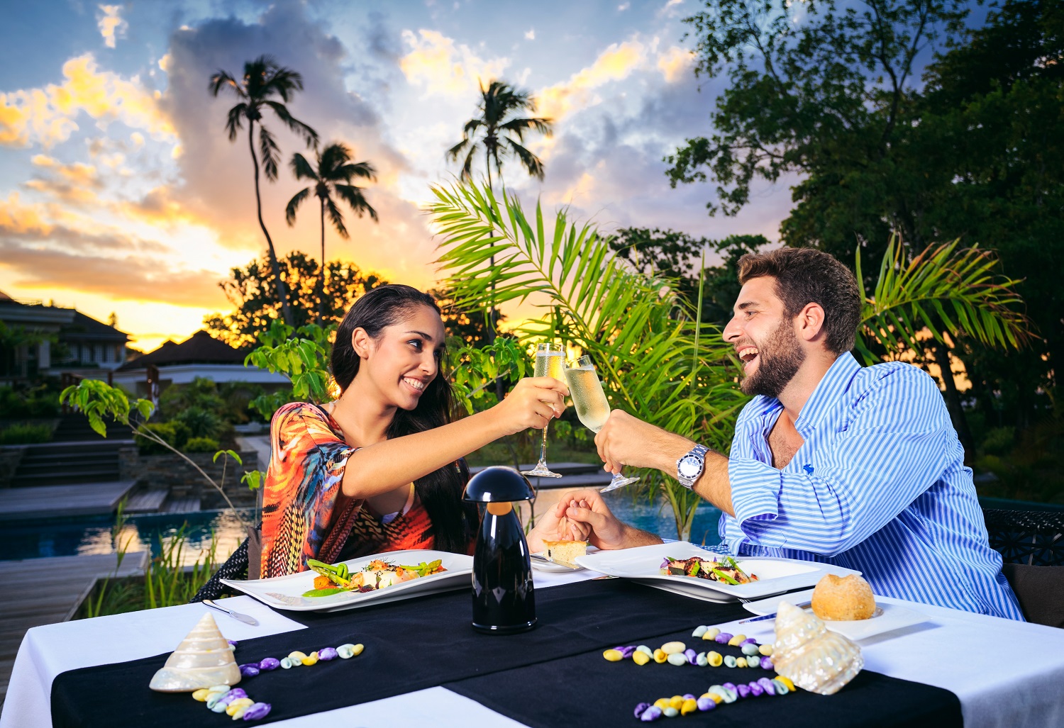 Couple toasting with champagne at a beachfront dinner table at sunset, palm trees and tropical vegetation surrounding them.