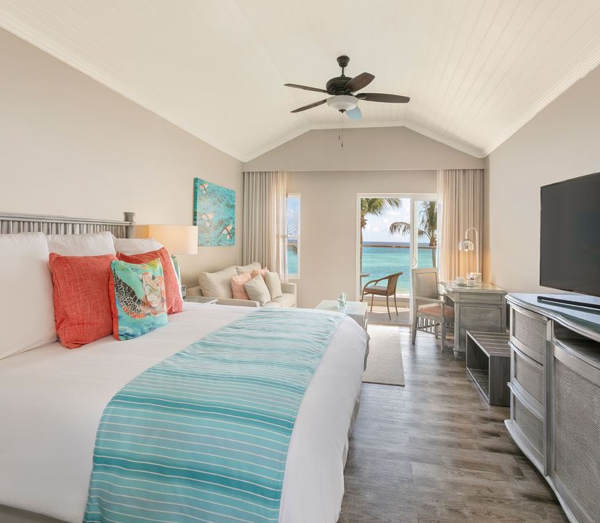 Bright beachfront bedroom with vaulted ceiling, queen bed with turquoise striped runner, coral pillows, ocean view through glass doors, and ceiling fan.