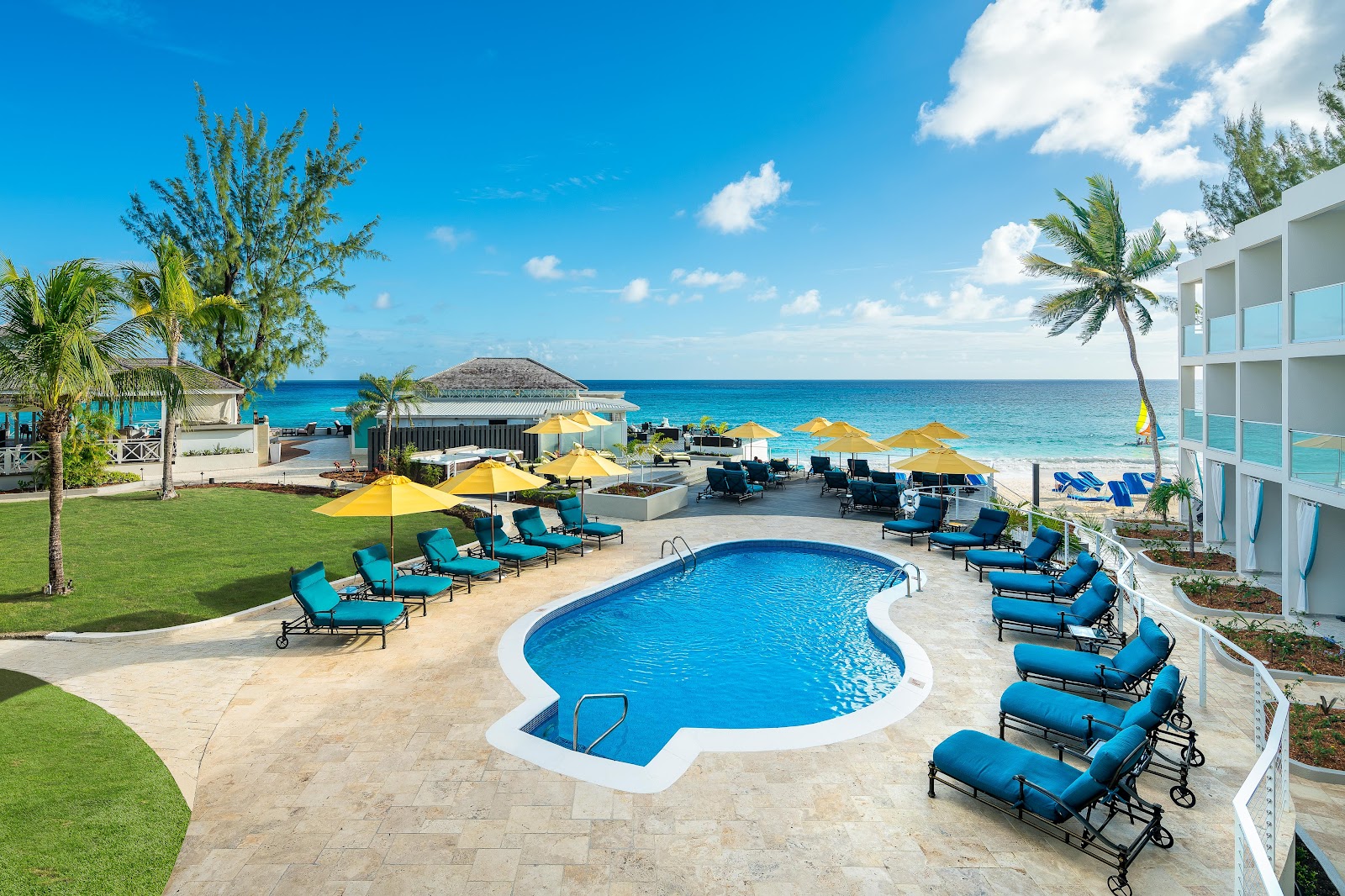Resort pool with blue loungers and yellow umbrellas, ocean and beach beyond, palm trees and white building at Sea Breeze Beach House.