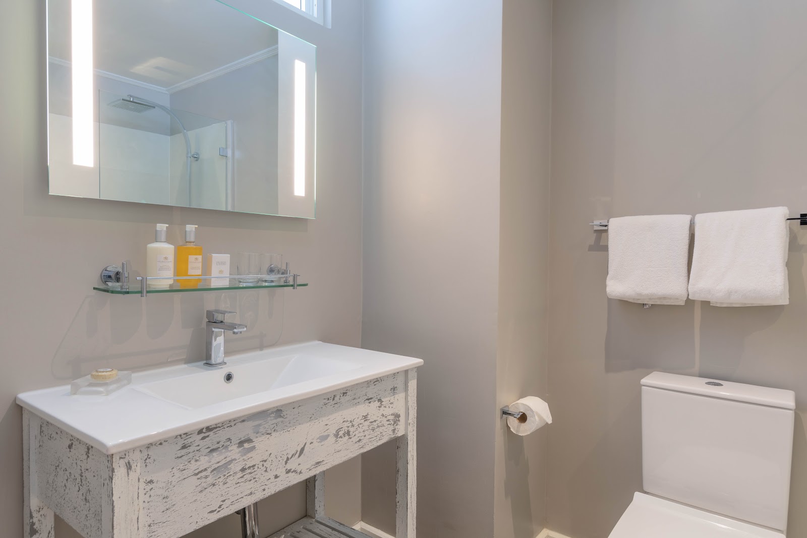 Modern bathroom with lit mirror cabinet, whitewashed vanity, glass shelf with toiletries, and folded white towels on rack.