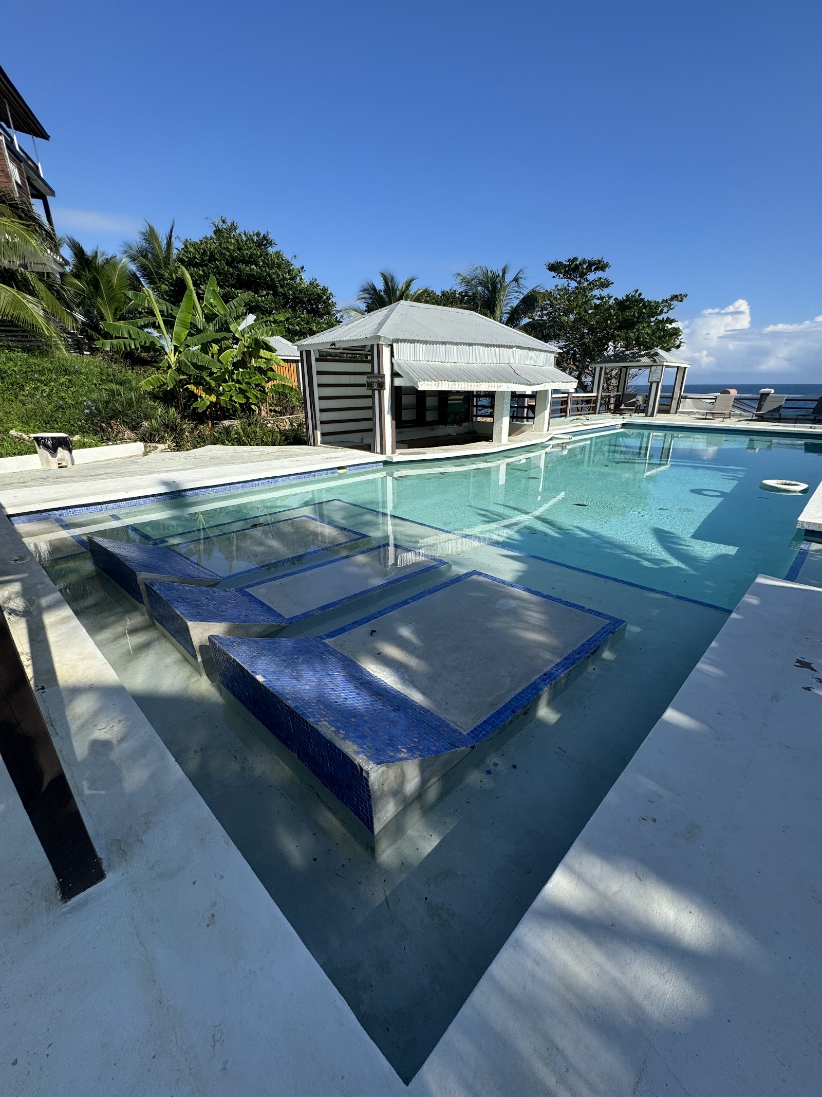 Swimming pool with submerged lounge platforms at Sea Cliff Resort & Spa, overlooking the ocean with a beach bar pavilion and tropical vegetation.