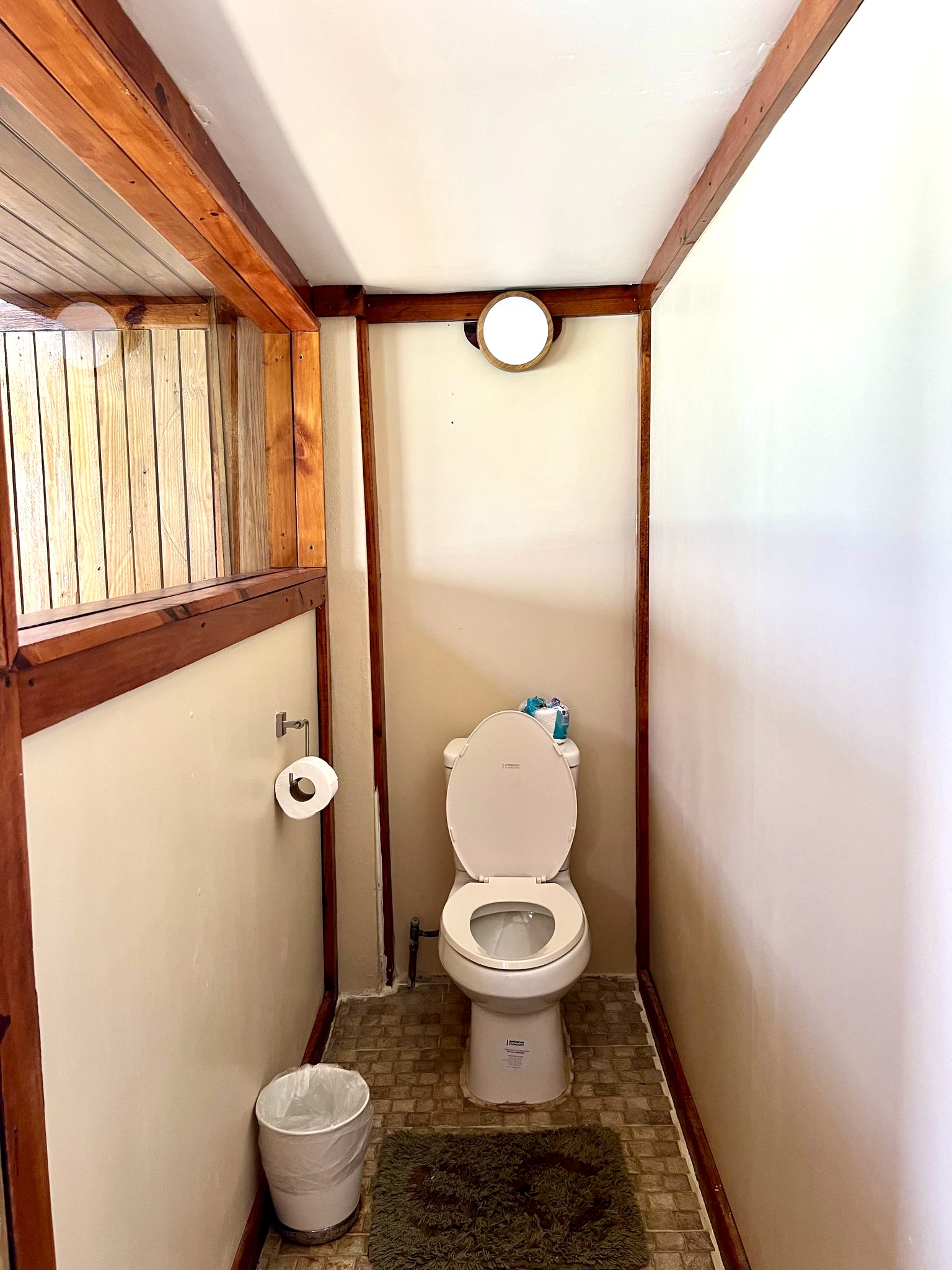 Bathroom interior with white toilet, cream walls, wooden beams, round mirror, and waste bin on brown tile floor.