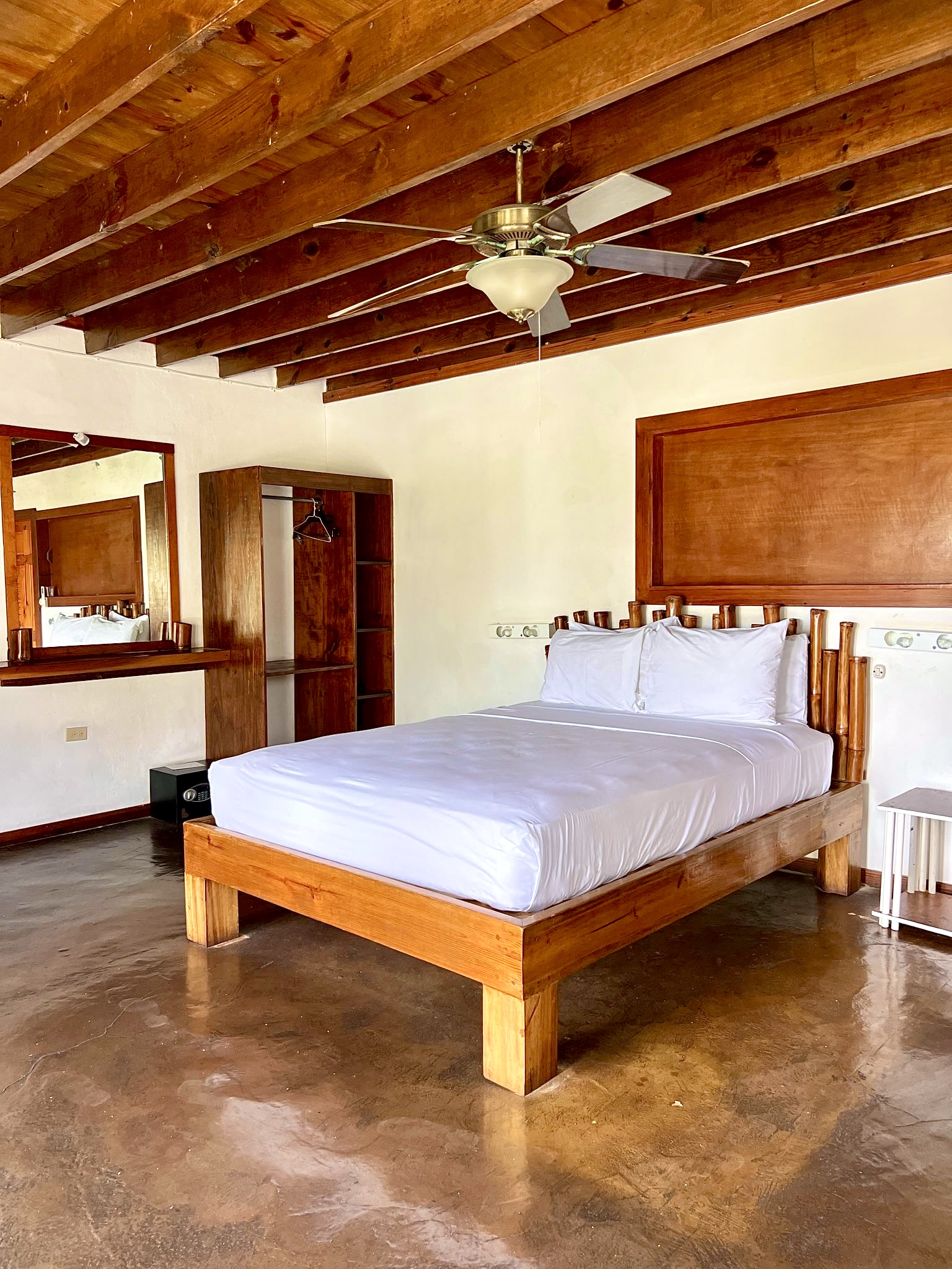 Spacious resort bedroom with wooden beam ceiling, queen bed with white linens, wooden furnishings, and concrete floor.