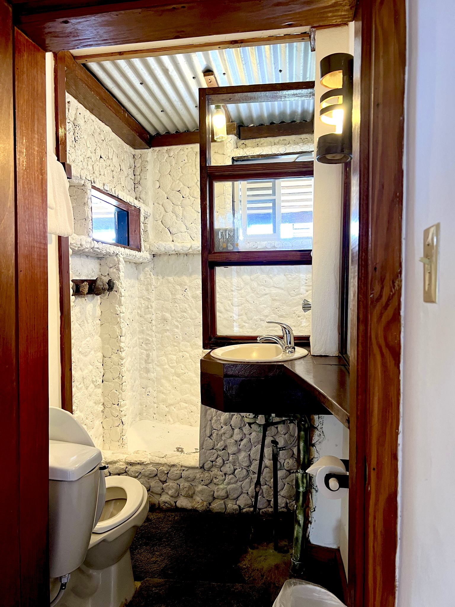 Bathroom at Sea Cliff Resort & Spa with white stone walls, wooden beams, round sink, toilet, and window overlooking the ocean.