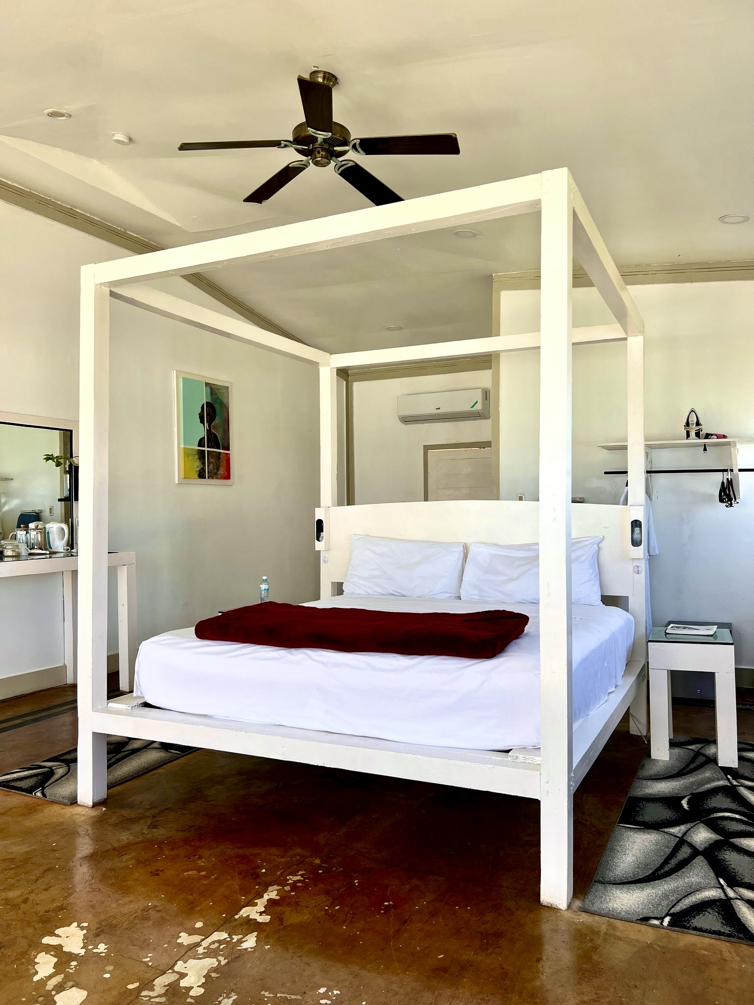 White four-poster bed with white pillows and dark red throw in a spacious room with ceiling fan, vanity desk, and polished concrete flooring.