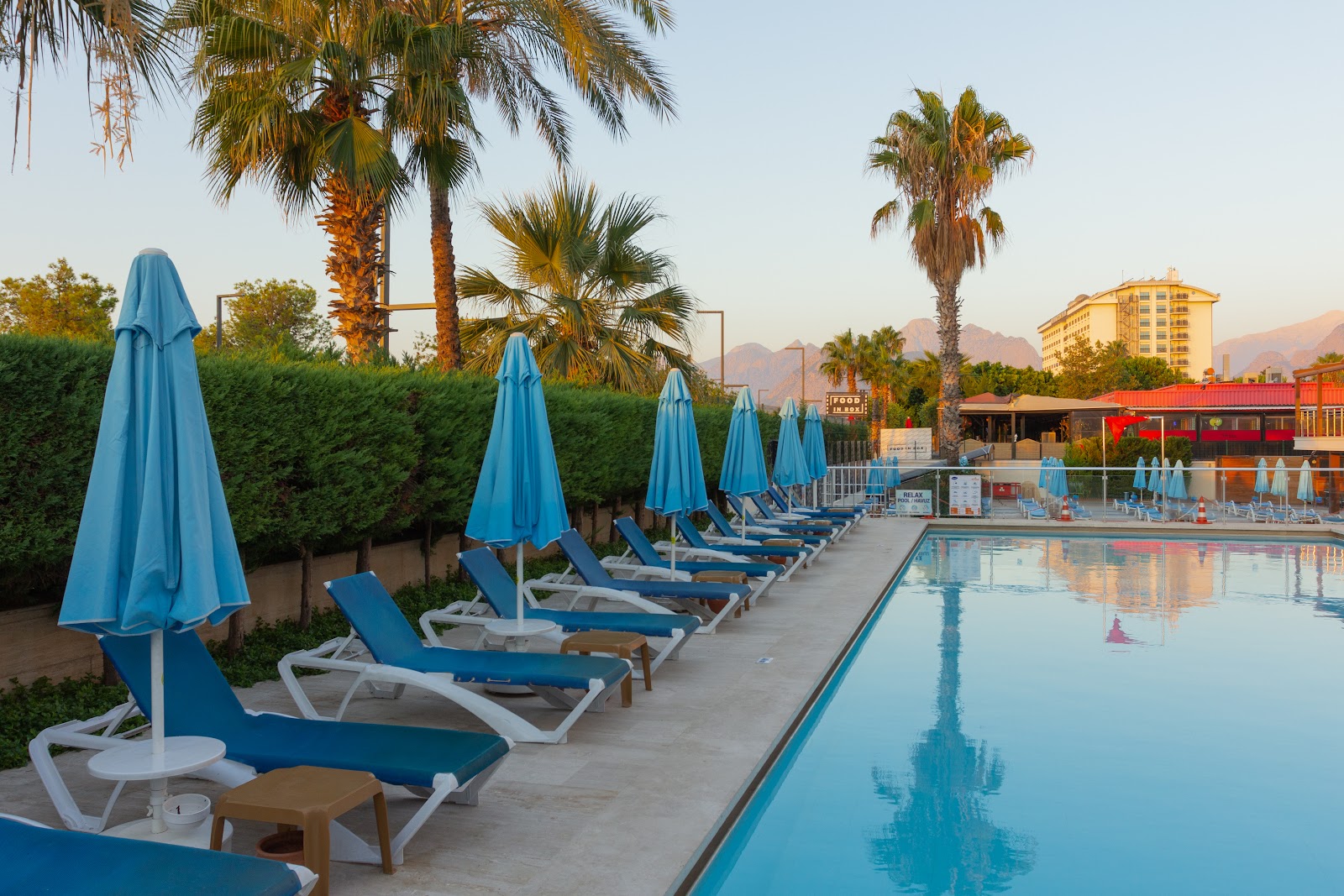 Blue loungers and closed umbrellas line the pool deck at Sealife Family Resort Hotel with palm trees and mountains in the background.
