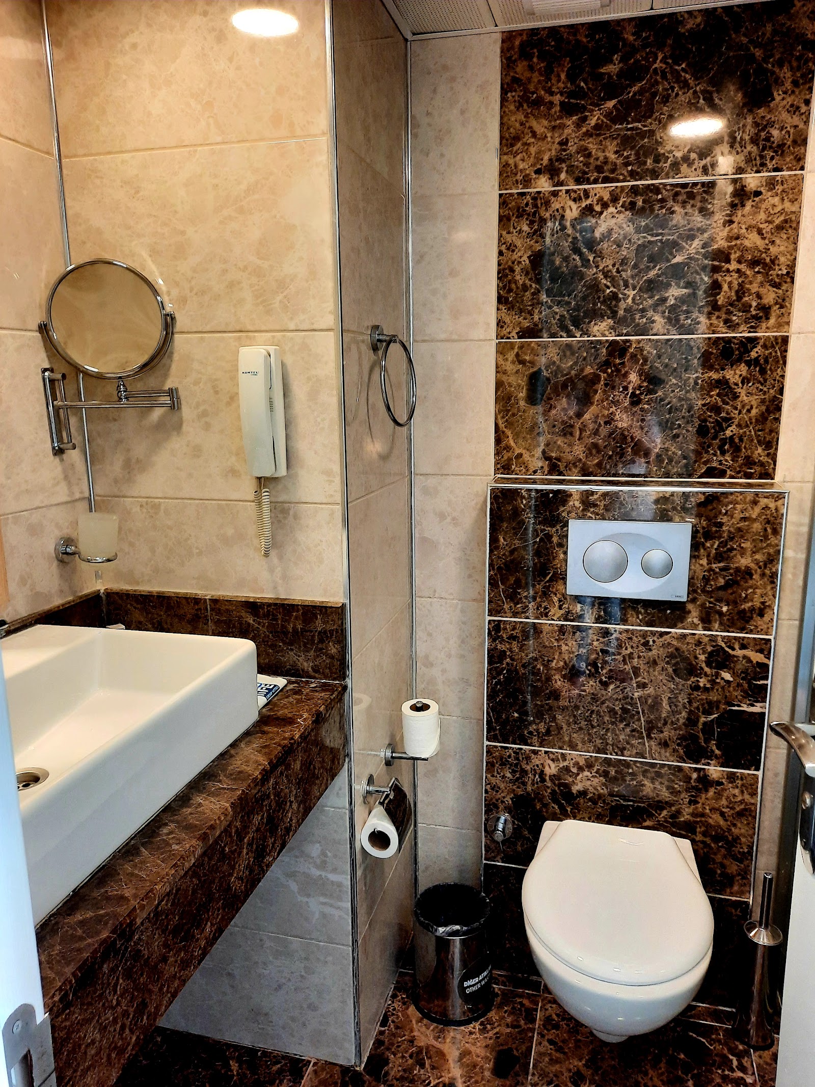 Modern bathroom with brown marble tiles, white rectangular sink, round mirror, walk-in shower, and wall-mounted toilet.