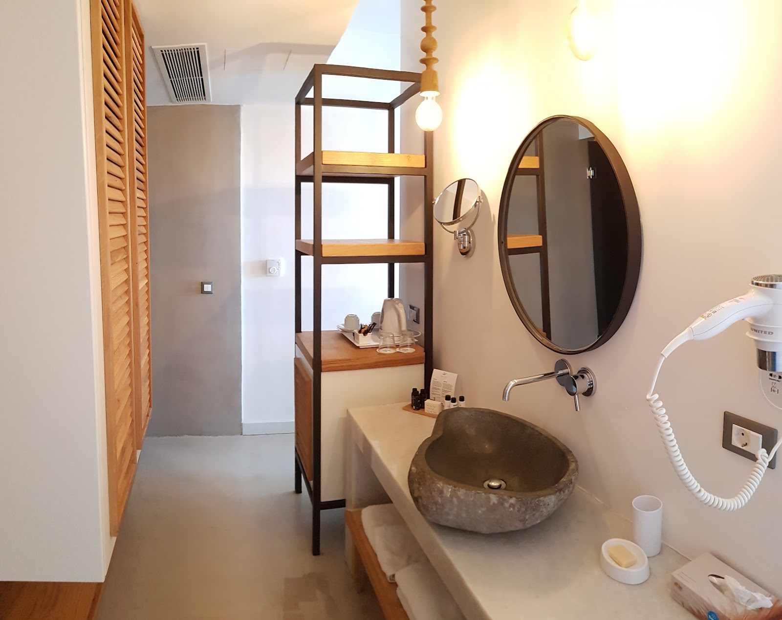 Bathroom vanity with round stone sink, circular mirror, and wooden shelving unit holding toiletries and supplies.