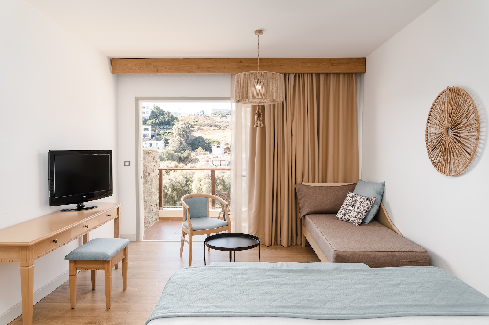 Clifftop resort with tan stone buildings overlooks a sandy beach with blue umbrellas and clear turquoise waters dotted with boats.