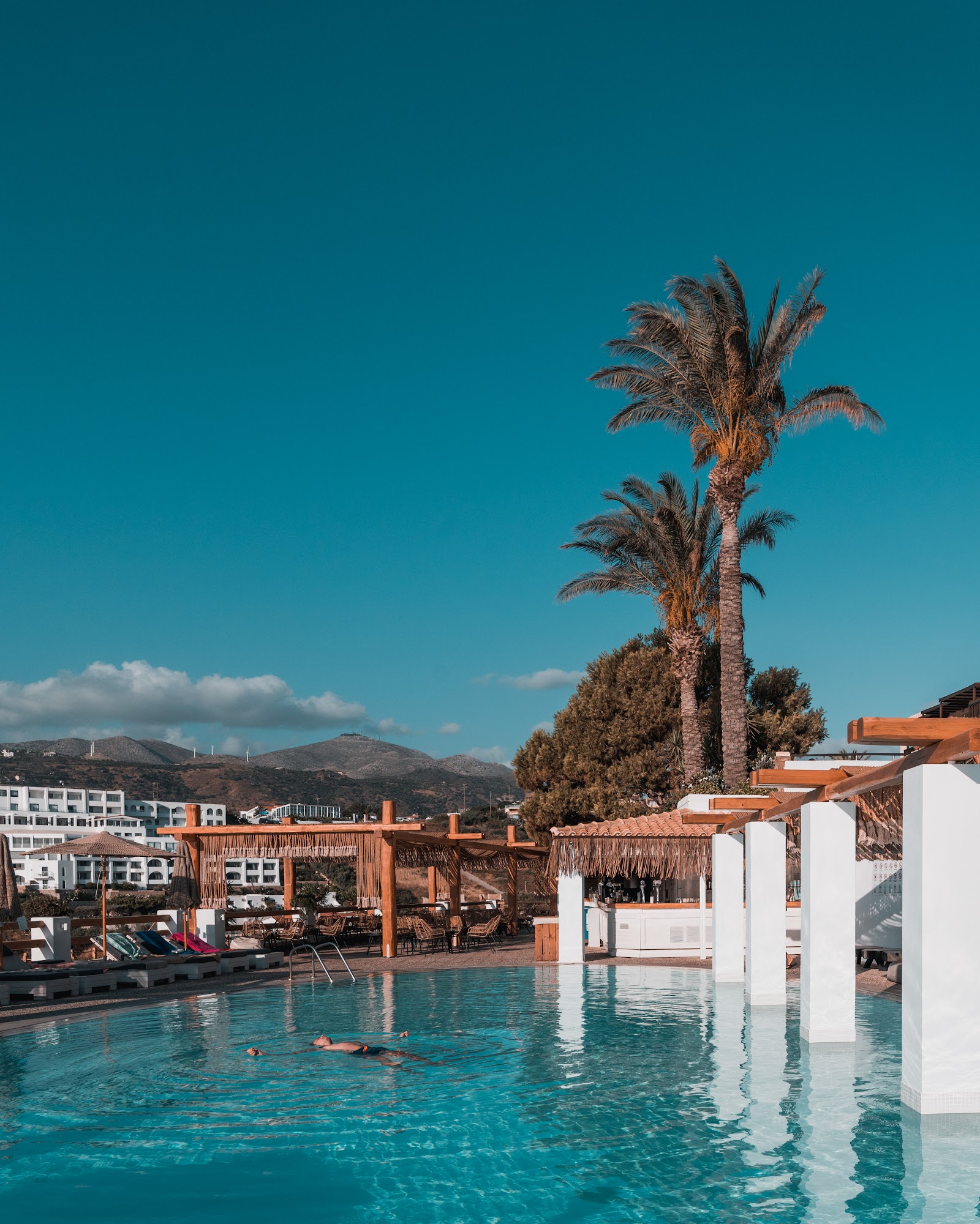 Beige resort courtyard with arched alcoves, wooden lounge chairs, central olive tree planter, and blue Mediterranean Sea and hillside visible beyond.