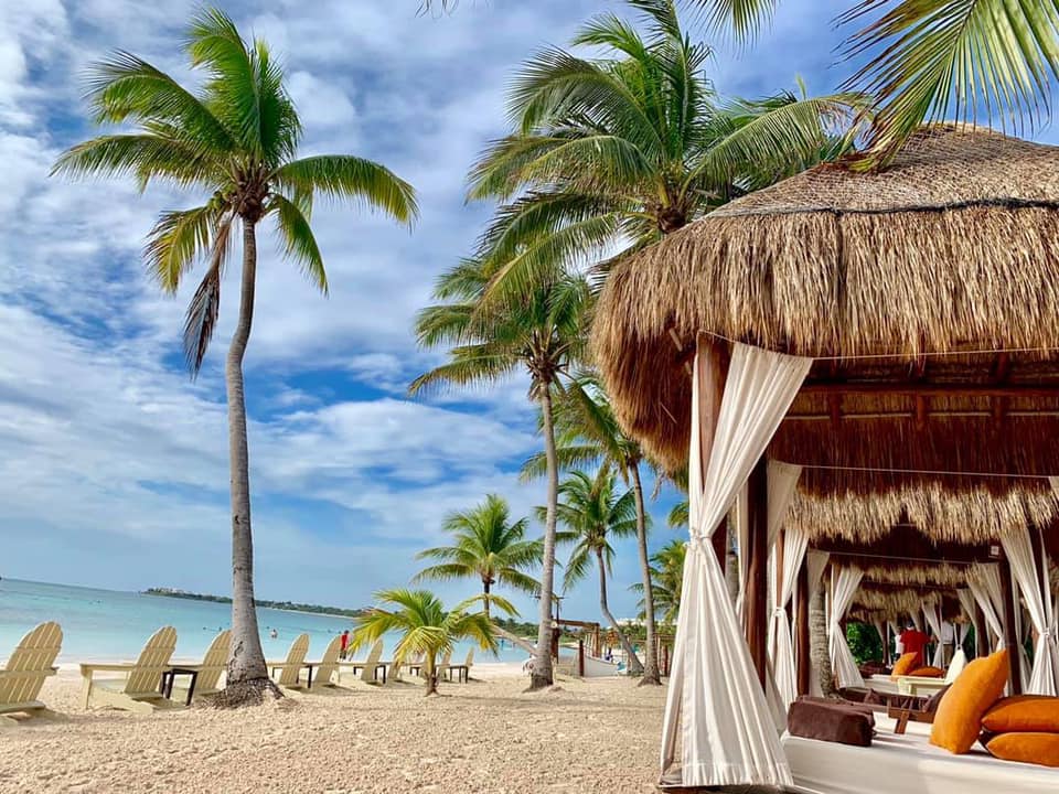 Beachfront cabanas with white curtains line the sand at Secrets Akumal Riviera Maya, with palm trees and turquoise water beyond.