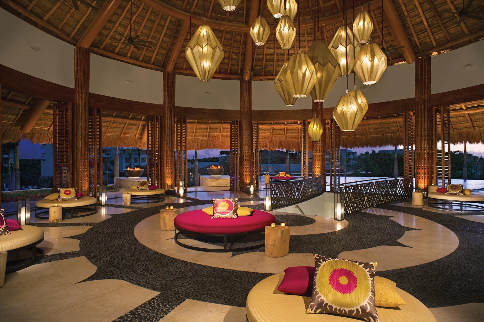 Circular lounge with thatched roof, gold pendant lights, red and yellow seating, and water views at dusk from Secrets Akumal Riviera Maya.