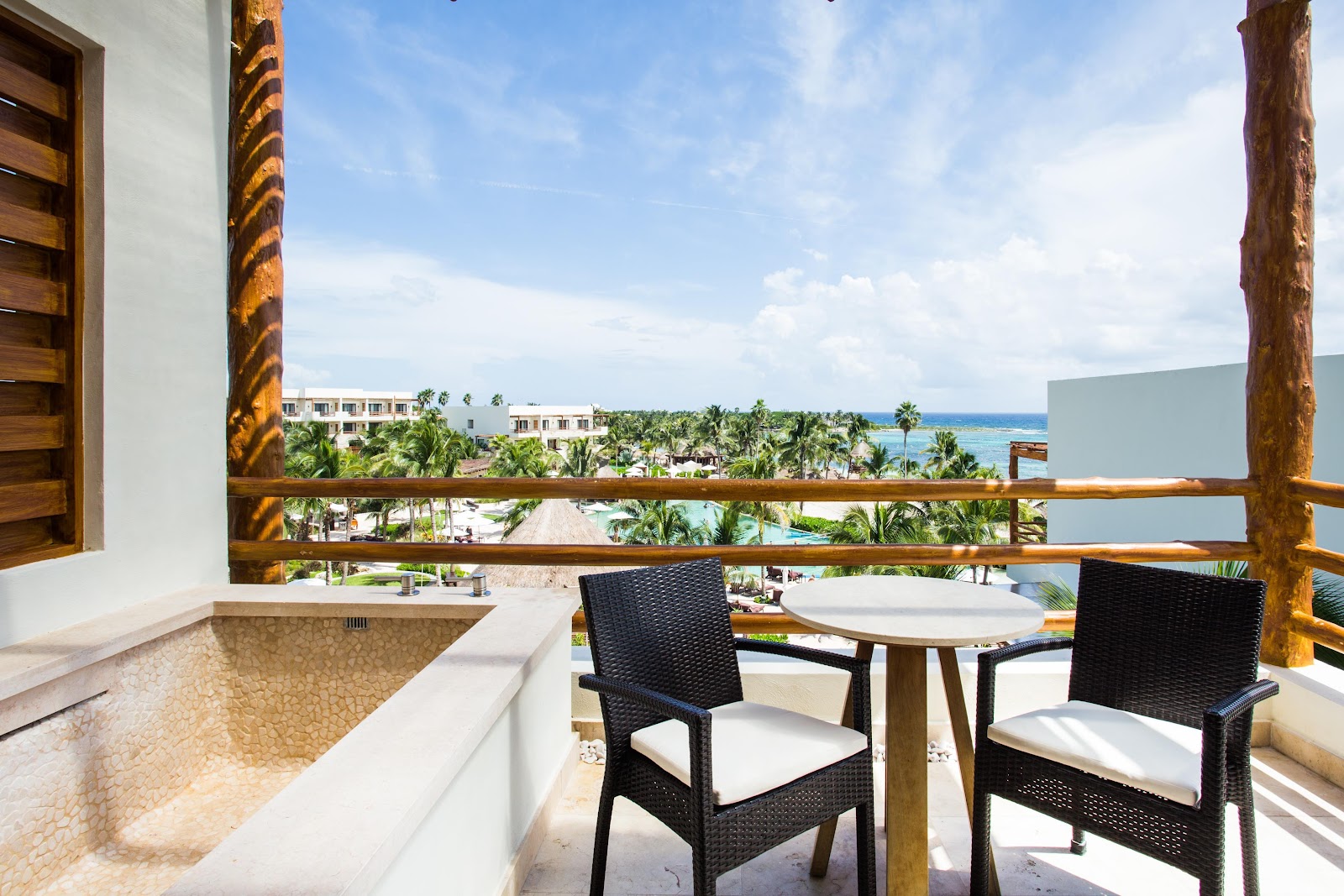 Balcony with two black wicker chairs at a round table overlooking palm trees, resort buildings, and turquoise ocean at Secrets Akumal Riviera Maya.