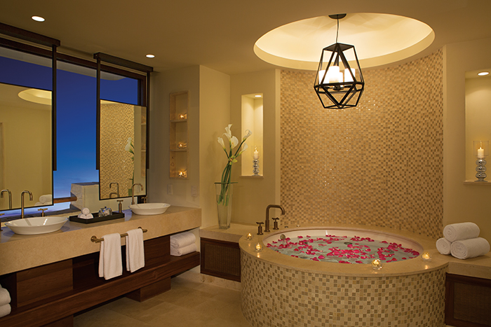 Luxury bathroom with soaking tub filled with red rose petals, dual vanity, geometric pendant light, and evening sky visible through windows.