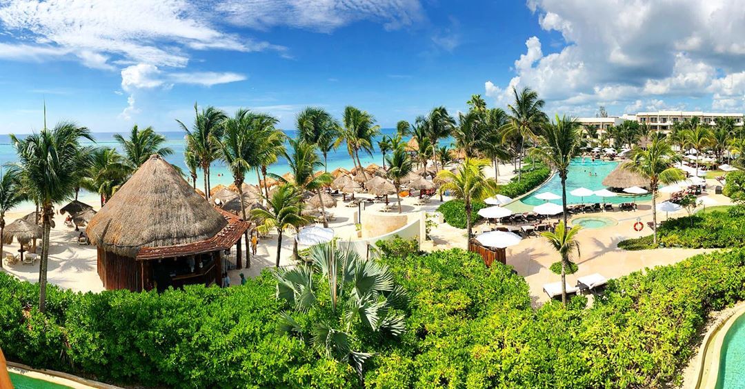 Secrets Akumal Riviera Maya beachfront with thatched-roof bar, palm trees, pool, and turquoise Caribbean Sea under blue sky.