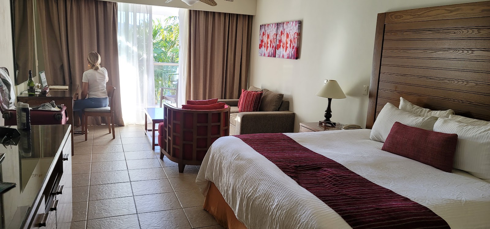 Spacious resort suite with queen bed, burgundy accents, sitting area, and balcony overlooking tropical grounds at Secrets Aura Cozumel.