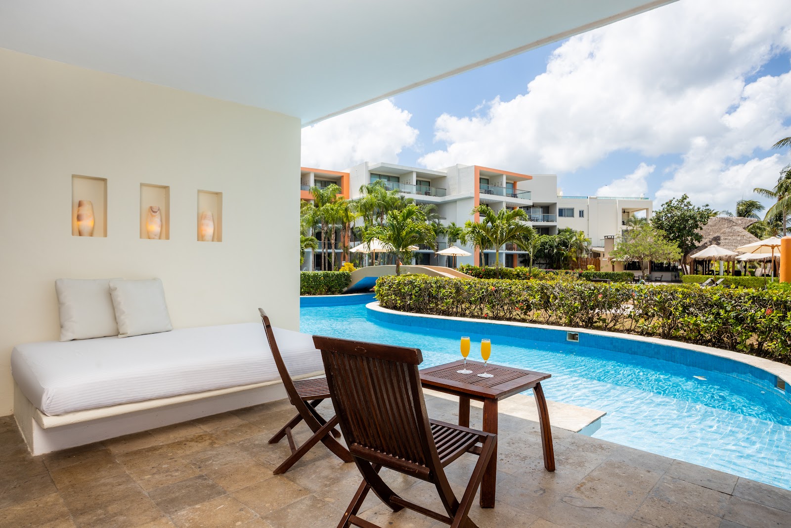 Covered patio with white daybed and wooden chair overlooking curved blue pool and modern resort buildings at Secrets Aura Cozumel.