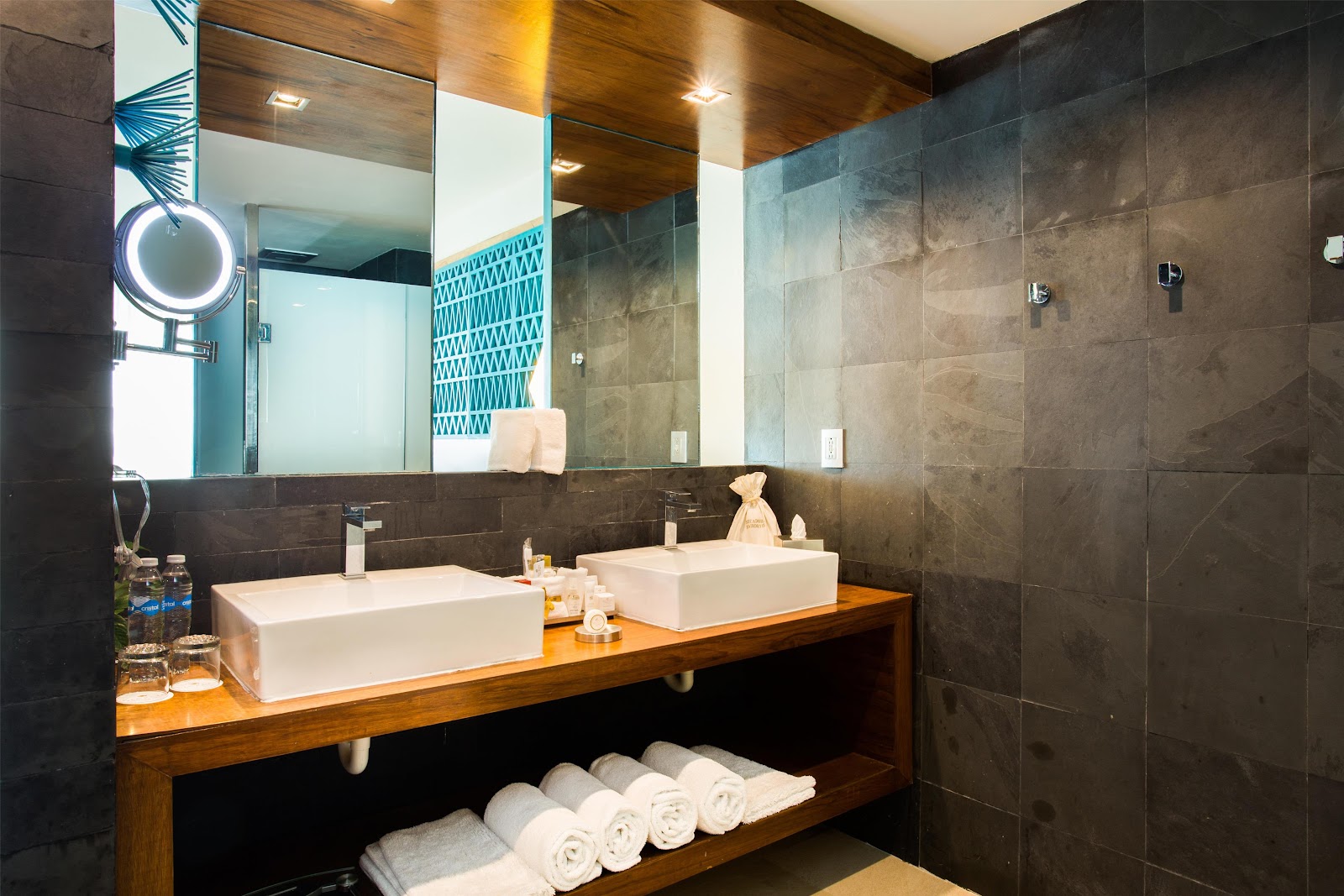 Modern bathroom at Secrets Aura Cozumel with dual white vessel sinks on wood vanity, dark tile walls, lit mirror, and rolled towels on shelf below.
