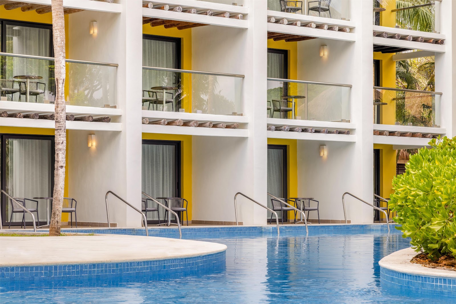Blue-tiled swimming pool with lounge chairs facing a white and yellow modern resort building with multiple balconies and palm trees.